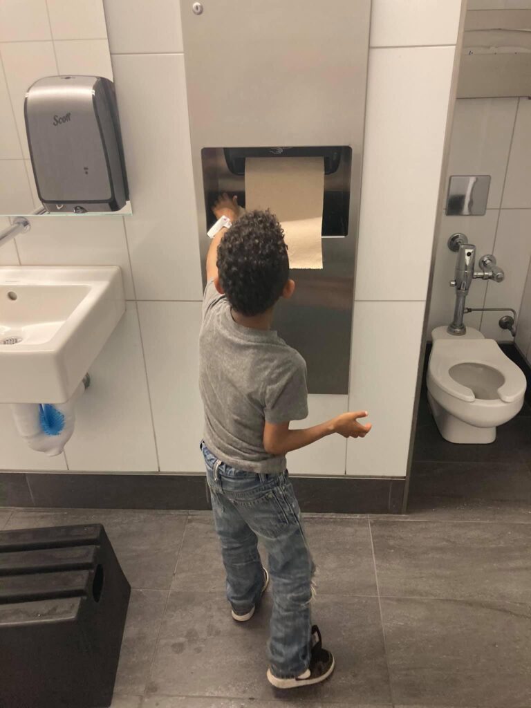 Child using an accessible bathroom with the sink, towel, and commode that are low to the ground.